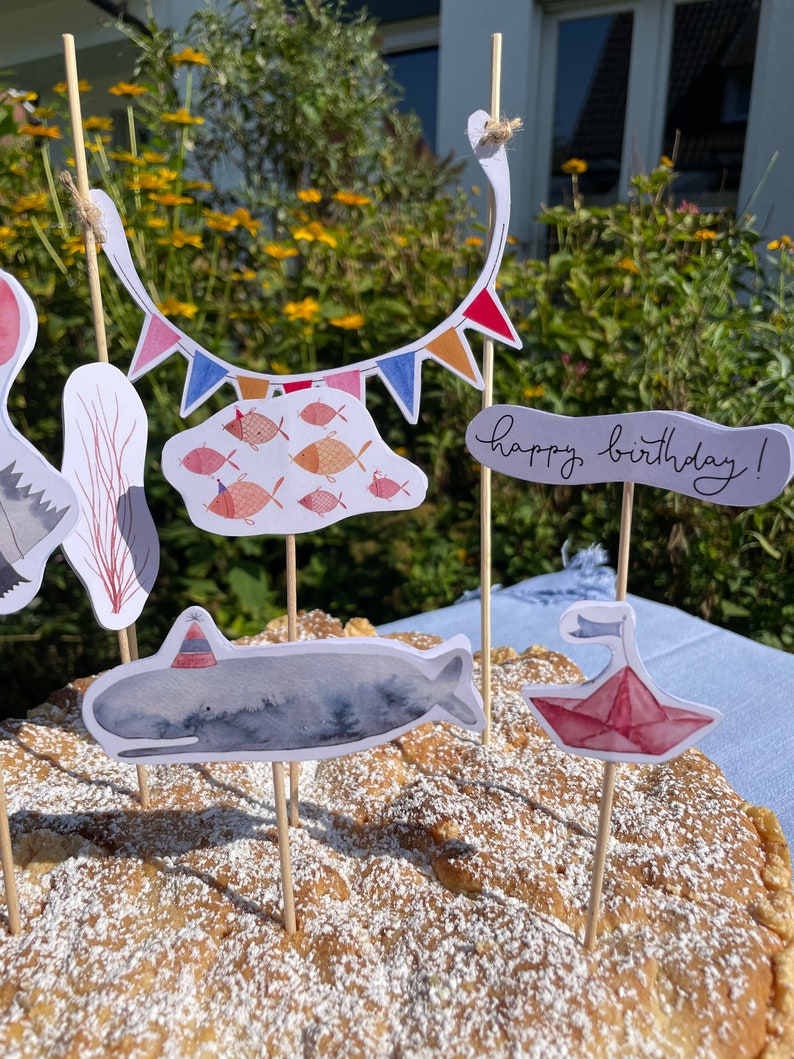 May include: A birthday cake decorated with a whale, a paper boat, and a string of colorful flags. The cake is topped with powdered sugar and the words "happy birthday!" are written on a banner.