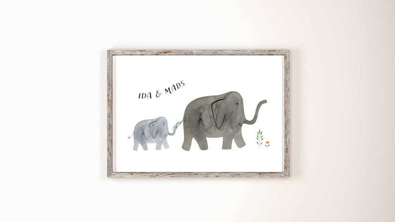 May include: Watercolor print of two gray elephants, one large and one small, connected by their trunks. The print is in a rustic wooden frame. Text at the top reads "IDA & MADS". Small green plants and a yellow flower are to the right of the elephants.
