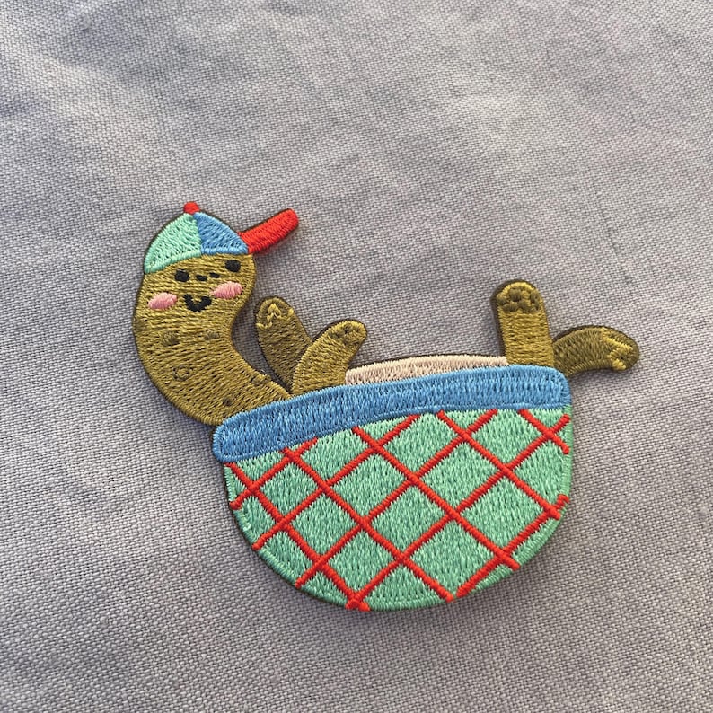 May include: Embroidered patch of a cartoon pickle in a baseball cap, lounging in a basket. The pickle is olive green with a smiling face and pink cheeks. The basket is blue and green with a red diamond pattern.