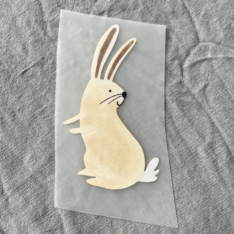 K&ouml;nnte beinhalten: Ein beigefarbener Hase mit braunen Ohren und einem wei&szlig;en Schwanz, aus einem Blatt durchsichtigem Papier ausgeschnitten.
