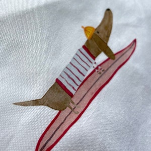 May include: A brown dachshund wearing a striped shirt and a yellow hat is riding a pink surfboard with red trim.