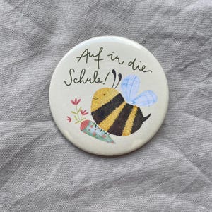 May include: A round button featuring a yellow and black striped bee with light blue wings. The bee is holding a floral-patterned cone. The text "Auf in die Schule!" is printed in black script above the bee on a cream-colored background.