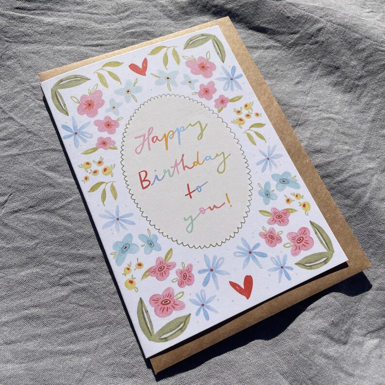Może przedstawiać: Kartka urodzinowa z kwiatowym wzorem i napisem "Happy Birthday to you!" w kolorowym skrypcie. Karta zawiera r&oacute;żowe, niebieskie i ż&oacute;łte kwiaty, zielone liście i czerwone serca. Jest umieszczona na brązowej kopercie.