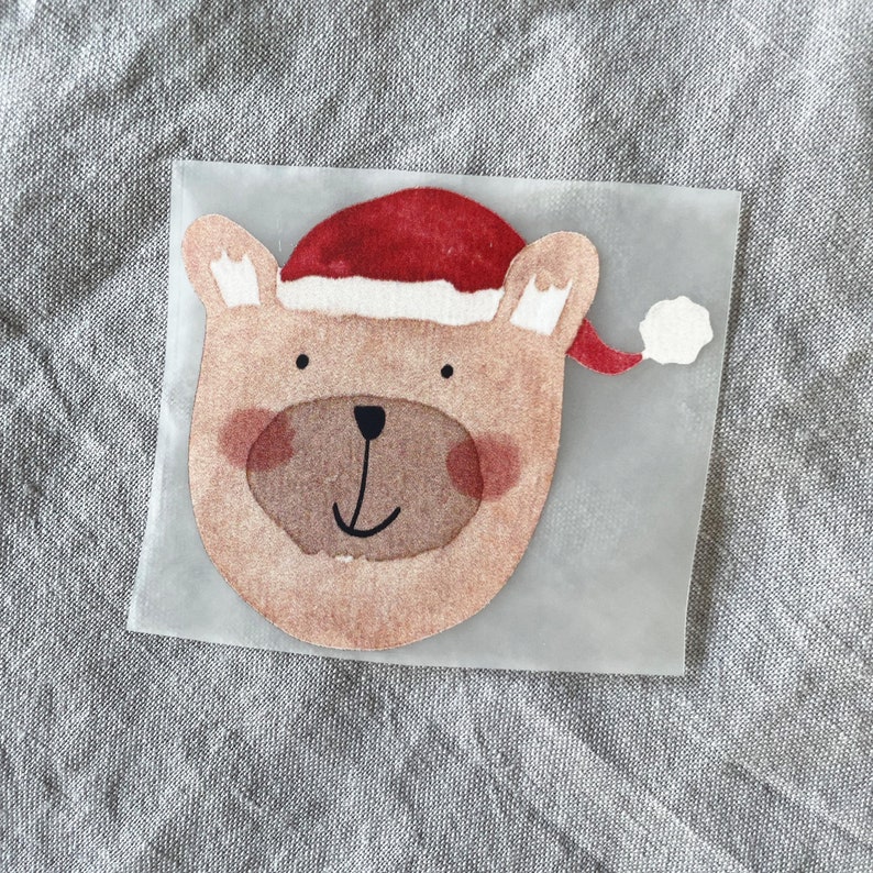 Op de afbeelding: Een bruine beer die een rode kerstmanmuts draagt met een witte pompon. De beer heeft een bruine neus en bruine ogen. De afbeelding is op een heldere achtergrond.