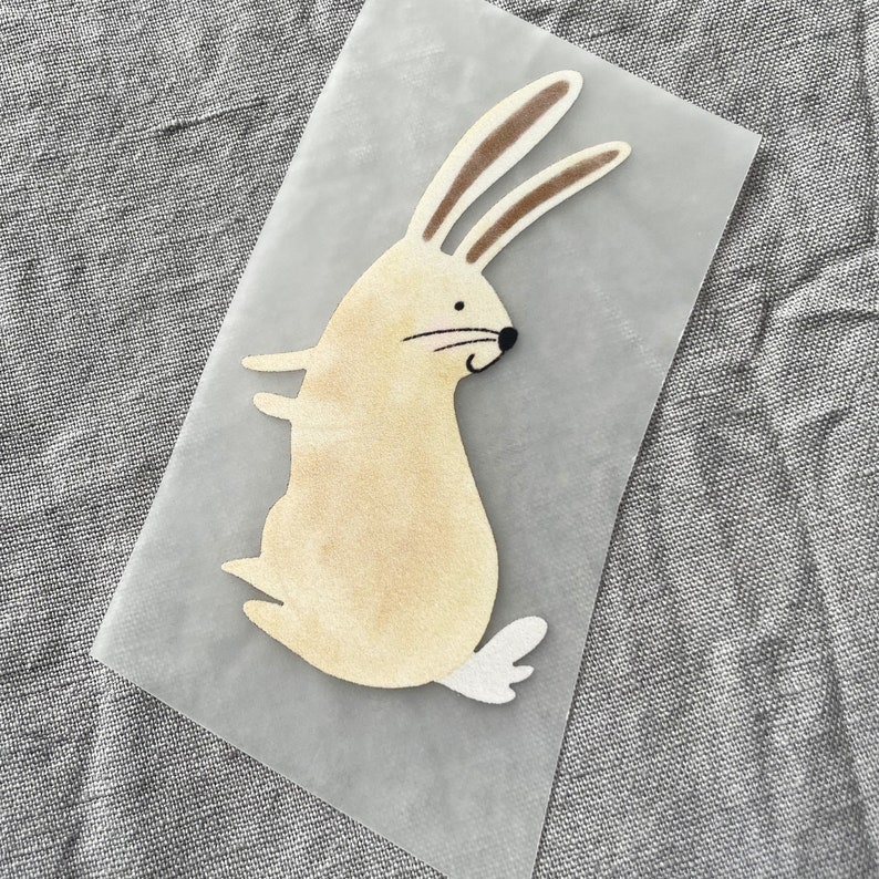 K&ouml;nnte beinhalten: Ein beigefarbener Filzhase mit braunen Ohren und einem wei&szlig;en Schwanz. Der Hase steht auf einer durchsichtigen Plastikfolie.