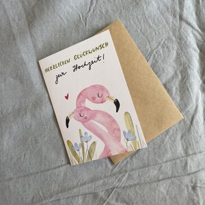 May include: A greetings card featuring a watercolour illustration of two pink flamingos with black beaks and a small red heart. The card has text in green and black, and is accompanied by a brown envelope.