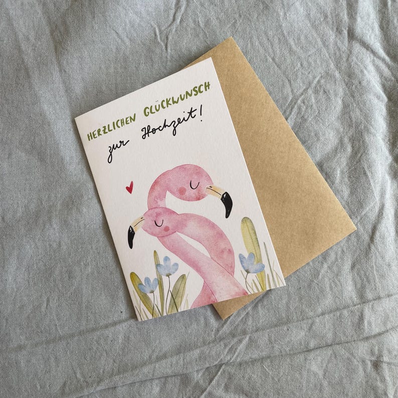 May include: A greetings card featuring a watercolour illustration of two pink flamingos with black beaks and a small red heart. The card has text in green and black, and is accompanied by a brown envelope.
