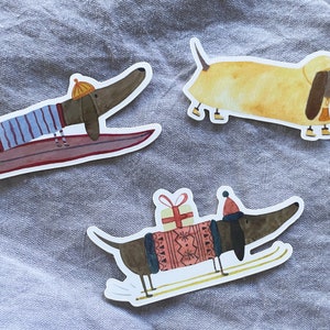 May include: Three dachshund dog stickers with different designs. One is surfing on a surfboard, another is wearing a yellow hat and a red and white striped sweater, and the third is wearing a red hat and a red and white sweater with a gift box on its back.