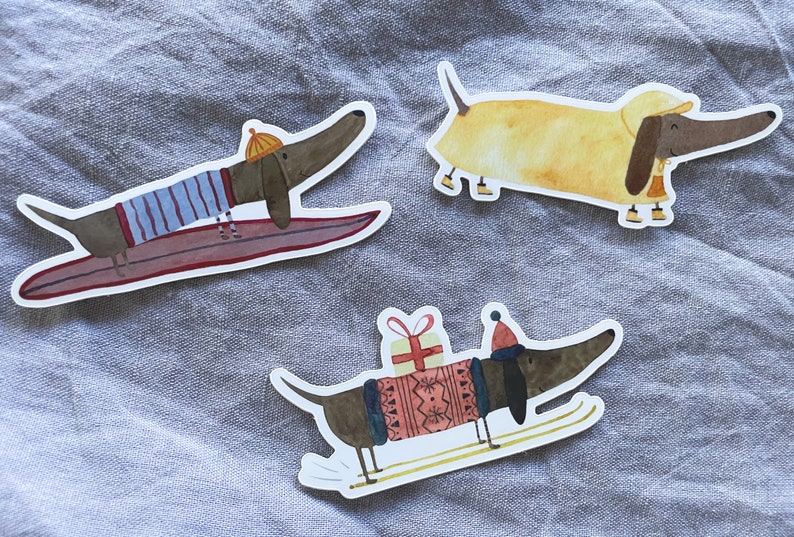 May include: Three dachshund dog stickers with different designs. One is surfing on a surfboard, another is wearing a yellow hat and a red and white striped sweater, and the third is wearing a red hat and a red and white sweater with a gift box on its back.