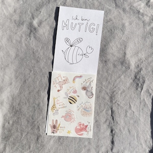 K&ouml;nnte beinhalten: Ein kleiner Notizblock mit den Worten "Ich bin MUTIG!" und einer Zeichnung einer Biene, die eine Blume h&auml;lt. Der Notizblock ist ge&ouml;ffnet und zeigt ein Blatt Aufkleber mit verschiedenen Illustrationen und deutschem Text.