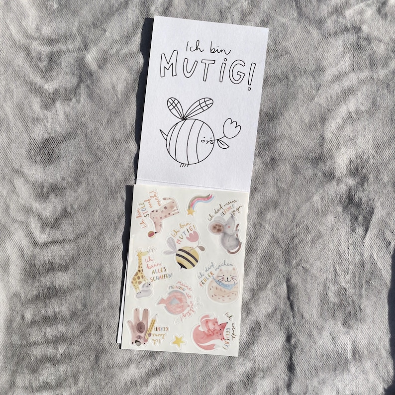 K&ouml;nnte beinhalten: Ein kleiner Notizblock mit den Worten "Ich bin MUTIG!" und einer Zeichnung einer Biene, die eine Blume h&auml;lt. Der Notizblock ist ge&ouml;ffnet und zeigt ein Blatt Aufkleber mit verschiedenen Illustrationen und deutschem Text.