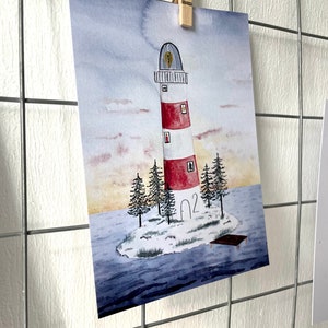 Puede incluir: Impresi&oacute;n en acuarela de un faro en una peque&ntilde;a isla. El faro es rojo y blanco con una parte superior negra y se encuentra en una peque&ntilde;a isla con &aacute;rboles de hoja perenne. La obra de arte est&aacute; sujeta a una rejilla de alambre con una pinza de madera.