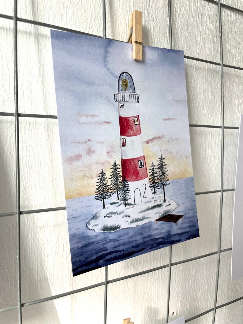 Puede incluir: Impresi&oacute;n en acuarela de un faro en una peque&ntilde;a isla. El faro es rojo y blanco con una parte superior negra y se encuentra en una peque&ntilde;a isla con &aacute;rboles de hoja perenne. La obra de arte est&aacute; sujeta a una rejilla de alambre con una pinza de madera.