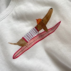 May include: A white t-shirt featuring a brown dachshund wearing a striped shirt and a yellow hat, riding a pink surfboard.