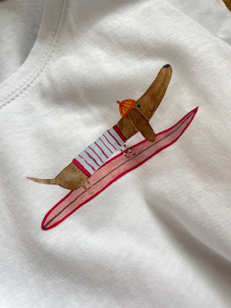 May include: A white t-shirt featuring a brown dachshund wearing a striped shirt and a yellow hat, riding a pink surfboard.