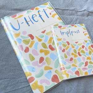 May include: Two clear plastic folders with a colorful abstract pattern. The larger folder has the text "U-Heft" on the front. The smaller folder has the text "Impfpass" on the front.