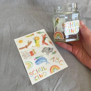 Pu&ograve; includere: Un barattolo di vetro trasparente con un adesivo che dice "SCHUL KIND" e un arcobaleno. Un foglio di adesivi colorati con animali, arcobaleni e le lettere "ABC" &egrave; accanto al barattolo. Gli adesivi sono in stile acquerello.