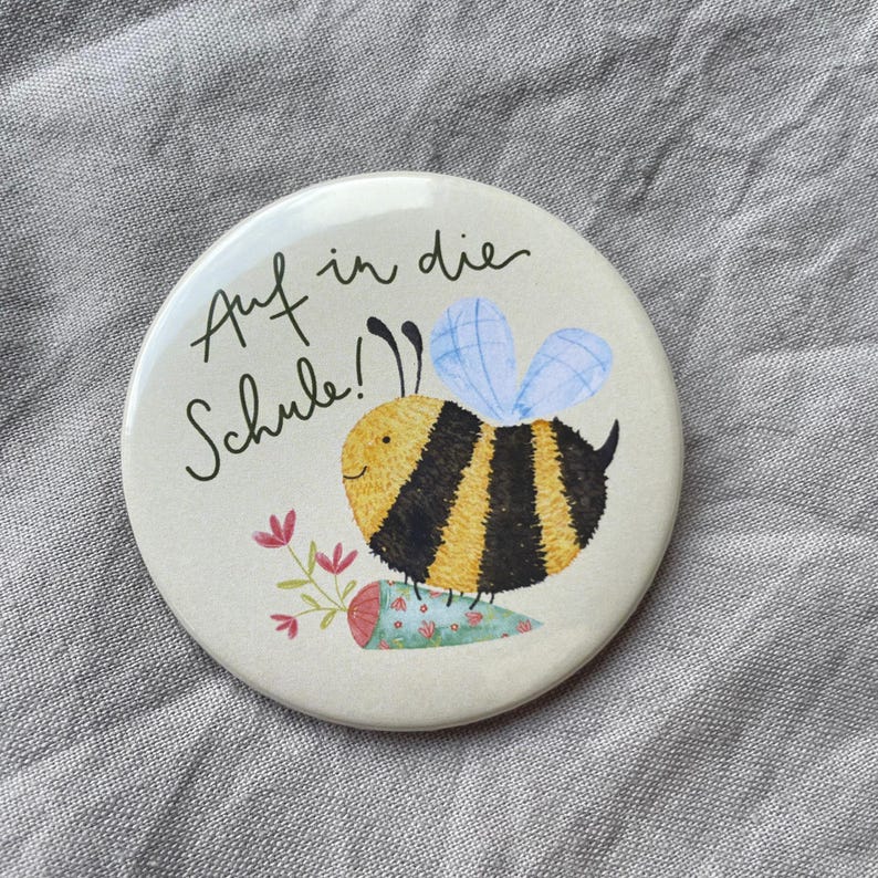May include: A round pinback button featuring a cream background with a black and yellow striped bee illustration. The bee holds a small floral cone. The text "Auf in die Schule!" is written in black cursive script above the bee illustration.