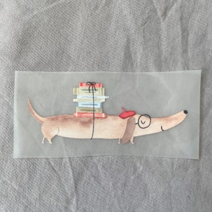 May include: A brown dachshund with a red beret and glasses is carrying a stack of books tied with a black ribbon.