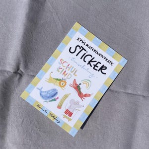 Pu&ograve; includere: Un foglio di adesivi con un bordo blu e giallo, con illustrazioni fantasiose di animali e un arcobaleno. La parola "STICKER" &egrave; ben visibile, insieme al testo "SCHUL KIND" e "Einschulung". Il foglio di adesivi &egrave; su una superficie grigia.