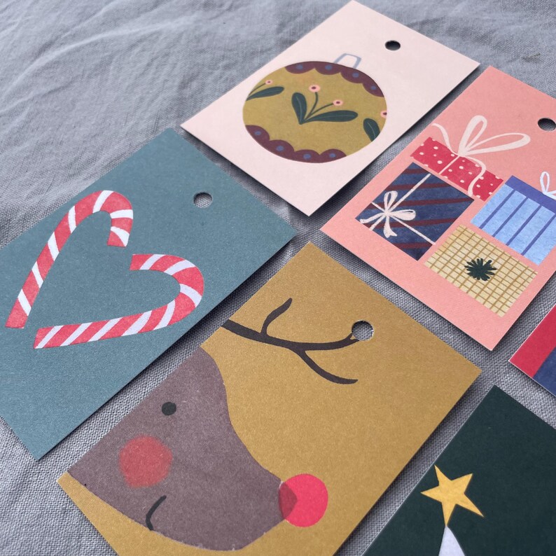 K&ouml;nnte beinhalten: Eine Sammlung von Weihnachtsgeschenkanh&auml;ngern mit verschiedenen festlichen Designs. Die Anh&auml;nger zeigen Illustrationen eines Zuckerstangenherzens, eines Rentiergesichts, verpackter Geschenke und einer Weihnachtskugel. Jeder Anh&auml;nger hat ein Loch zum Befestigen an Geschenken.