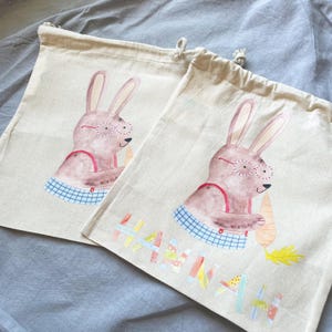 May include: Two beige canvas bags with drawstring closures. Each bag features a hand-painted illustration of a brown bunny wearing red glasses and holding a carrot. The bag on the right has the name "Hannah" written in colorful letters.