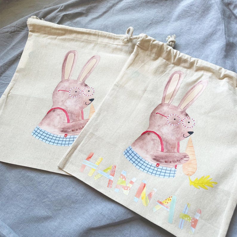 May include: Two beige canvas bags with drawstring closures. Each bag features a hand-painted illustration of a brown bunny wearing red glasses and holding a carrot. The bag on the right has the name "Hannah" written in colorful letters.
