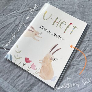 May include: A personalized U-Heft book cover featuring watercolor illustrations of two bunnies, flowers, and foliage. The cover includes the name "Leonie M&uuml;ller" in handwritten script, with the title "U-Heft" in colorful lettering. The words "Personalizierung von Hand" and "im Handlettering Stil" are also visible.