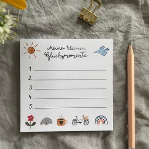 Op de afbeelding: Een wit notitieblok met de tekst "Meine kleinen Gl&uuml;cksmomente" en genummerde lijnen. Het notitieblok bevat aquarel illustraties van een zon, vogel, bloem, hond, koffiekopje, fiets en regenboog. Een potlood en gouden paperclip zijn ook aanwezig.