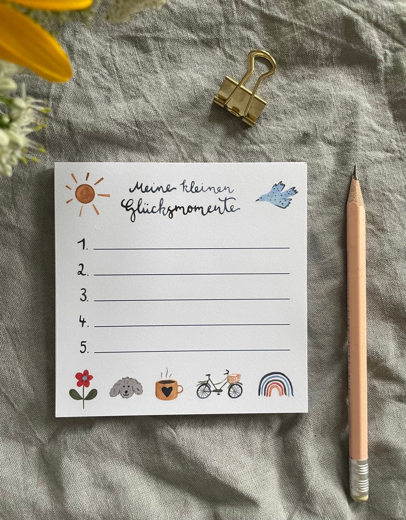 Op de afbeelding: Een wit notitieblok met de tekst "Meine kleinen Gl&uuml;cksmomente" en genummerde lijnen. Het notitieblok bevat aquarel illustraties van een zon, vogel, bloem, hond, koffiekopje, fiets en regenboog. Een potlood en gouden paperclip zijn ook aanwezig.