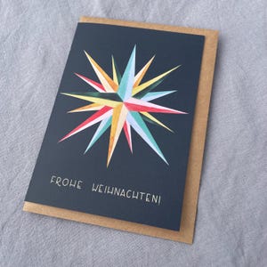 May include: Greeting card with a navy blue background and a colorful starburst design. The starburst features red, yellow, green, and blue hues. The card is paired with a brown envelope, and the text "FROHE WEIHNACHTEN!" is printed on the front.