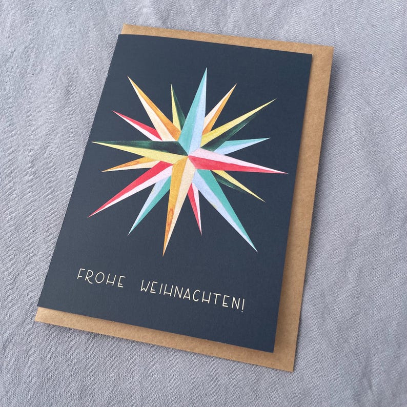 May include: Greeting card with a navy blue background and a colorful starburst design. The starburst features red, yellow, green, and blue hues. The card is paired with a brown envelope, and the text "FROHE WEIHNACHTEN!" is printed on the front.