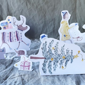 May include: A set of paper Christmas decorations featuring a reindeer wearing a sweater, a Christmas tree with lights, a house, a snowman, a hedgehog, a girl in a yellow coat, a boy in a blue shirt, and a pink cat.