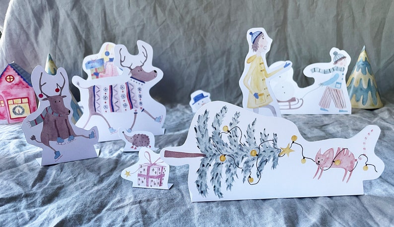 May include: A set of paper Christmas decorations featuring a reindeer wearing a sweater, a Christmas tree with lights, a house, a snowman, a hedgehog, a girl in a yellow coat, a boy in a blue shirt, and a pink cat.