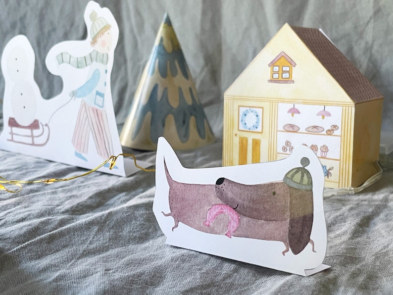 May include: A set of paper cutouts featuring a snowman, a person pulling a sled, a Christmas tree, a house with a bakery inside, and a dachshund wearing a hat and holding a pink donut.