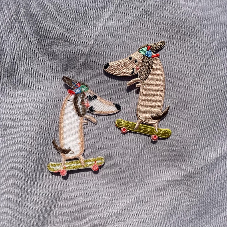 May include: Two embroidered patches featuring dachshund dogs on skateboards. The dogs are tan and white with black accents, wearing colorful hats. The skateboards are yellow-green with red wheels. The patches are on a gray fabric background.