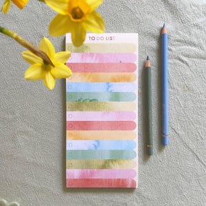 Può includere: Un elenco di cose da fare ad acquerello con 10 righe colorate e caselle di controllo. L'elenco è su uno sfondo bianco con due matite colorate, una blu e una grigia, che giacciono sulla superficie.