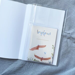 May include: A clear plastic vaccine passport holder with a white interior. The passport inside has the word "Impfpass" in blue script, and features two dachshund illustrations with hats. The background is a light grey fabric.