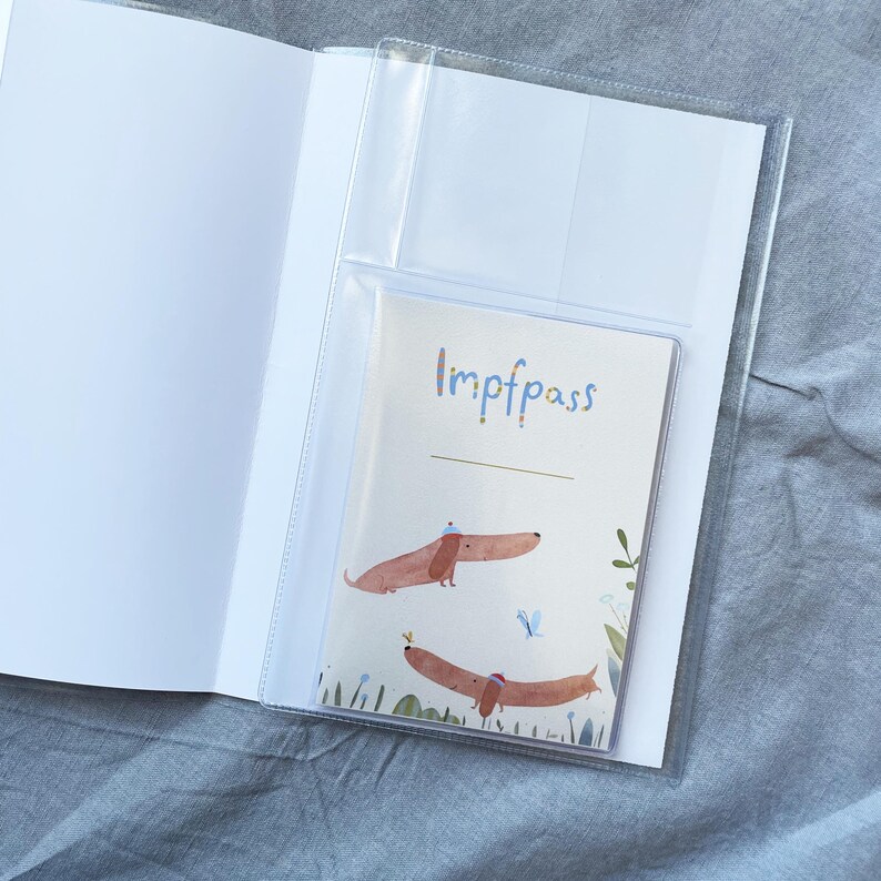 May include: A clear plastic vaccine passport holder with a white interior. The passport inside has the word "Impfpass" in blue script, and features two dachshund illustrations with hats. The background is a light grey fabric.