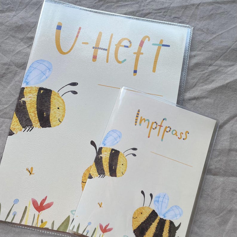 May include: Two protective covers for documents, one labeled U-Heft and the other Impfpass. Both feature a cream background with illustrations of yellow and black striped bees, blue wings, and small colorful flowers at the bottom edge.