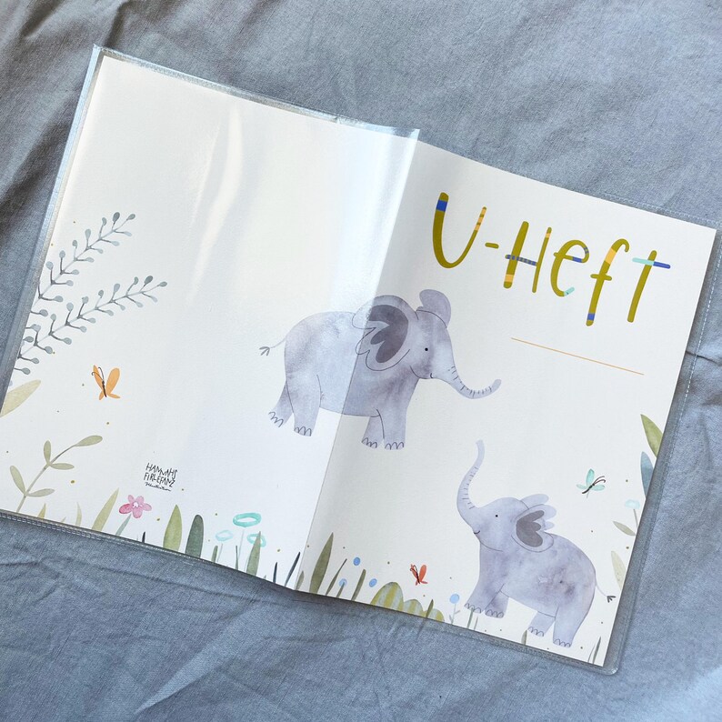 May include: A white book with a clear plastic cover. The front cover features a watercolour illustration of two elephants in a field of flowers and butterflies. The text "U-Heft" is written in gold letters on the cover.