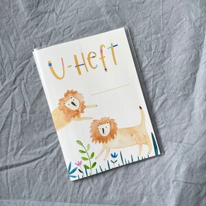K&ouml;nnte beinhalten: Ein wei&szlig;es Heft mit dem Text "U-Heft" in Blau und Gelb. Der Umschlag zeigt eine Aquarell-Illustration von zwei L&ouml;wen in einem grasbewachsenen Feld.