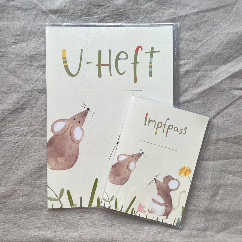May include: A cream-colored U-Heft booklet and a smaller Impfpass booklet, both protected by clear plastic covers. Each booklet features watercolor illustrations of brown mice, green grass, and a yellow dandelion, with text printed in green.