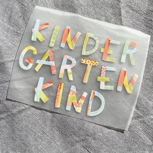 Peut inclure: Une feuille de plastique transparente avec les mots "Kindergarten Kind" &eacute;crits en lettres color&eacute;es et &agrave; motifs. Les lettres sont en papier et ont un aspect artisanal et fantaisiste.