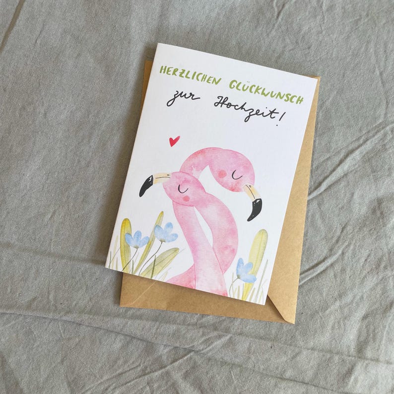 May include: A wedding greeting card featuring a watercolour illustration of two pink flamingos with black beaks and a red heart. The card has the text "HERZLICHEN GL&Uuml;CKWUNSCH zur Hochzeit!" in green and black. The card is inside a brown envelope.