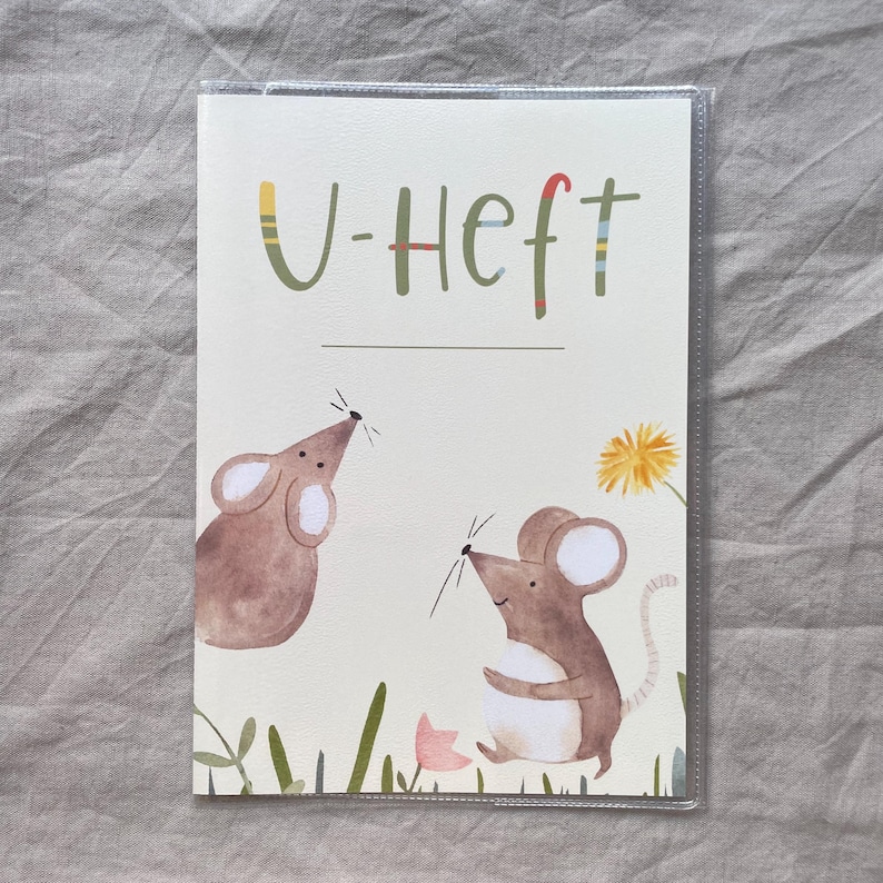 May include: A rectangular booklet cover with a cream background featuring the text U-Heft in green letters. The illustration shows two brown mice, a yellow dandelion, and a small pink flower, all enclosed in a clear protective plastic sleeve.