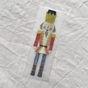 Può includere: Illustrazione ad acquerello di un soldato schiaccianoci. La figura indossa una giacca rossa e oro, pantaloni blu e stivali neri. Una corona d'oro è sulla testa dello schiaccianoci. L'illustrazione è su sfondo trasparente.