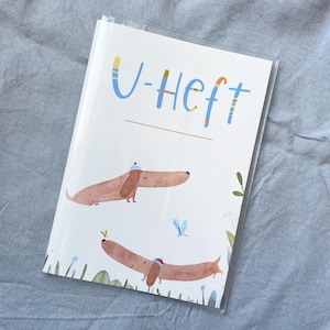 May include: A white notebook with the text "U-Heft" in blue and yellow lettering. The cover features illustrations of two brown dachshunds wearing hats, a blue butterfly, and green foliage.