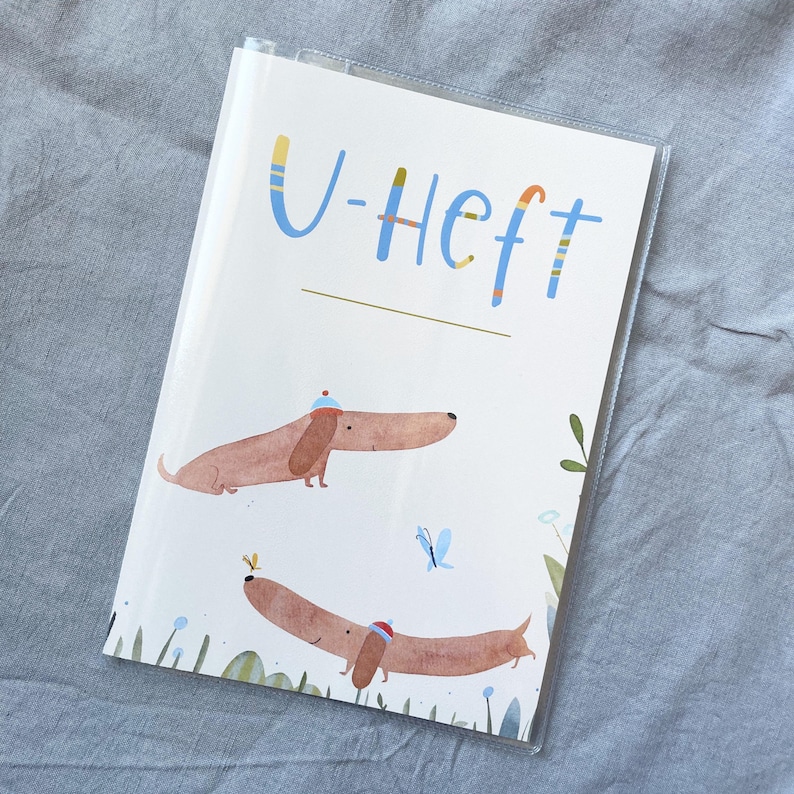 May include: A white notebook with the text "U-Heft" in blue and yellow lettering. The cover features illustrations of two brown dachshunds wearing hats, a blue butterfly, and green foliage.