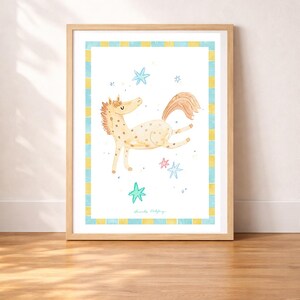 May include: A framed watercolor print of a whimsical horse with a light brown mane and tail, and a spotted body. The horse is depicted mid-leap with stars scattered around it. The print has a blue and yellow border and is set in a wooden frame.